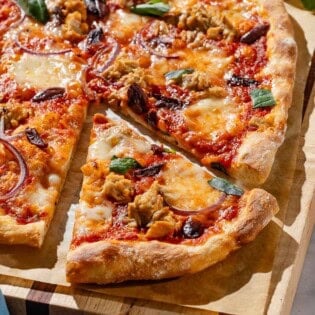 A close up photo of tuna pizza on a piece of parchment paper on a wooden cutting board, with one slice cut. Next to this is a cloth napkin and a bowl of basil.